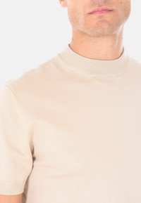Uomo che indossa una maglietta a maniche corte in maglia beige chiaro con colletto alla coreana su uno sfondo semplice.