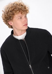 Jaunas vyras su garbanotais blondiniais plaukais vilki juodą zip-up striukę virš baltos marškinėlių, žiūrėdamas į kairę prieš baltą foną.