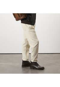 Pantalon en velours côtelé beige avec une coupe droite et une texture visible, associé à des bottes marron foncé à lacets. Sac à dos en cuir marron porté sur l'épaule.