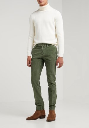 Mann trägt weißen Rollkragenpullover, olivgrüne Slim-Fit-Hose und braune Wildleder-Stiefeletten, steht vor unifarbenem Hintergrund.