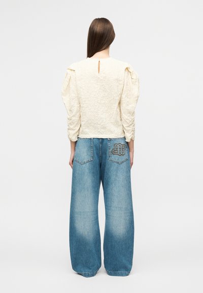 Φωτεινό μπεζ τοπ με υφή και φουσκωτά μανίκια, με άνοιγμα στην πλάτη. Μπλε τζιν παντελόνι με φαρδιά μπατζάκια και ραμμένο σχέδιο στην πίσω τσέπη.