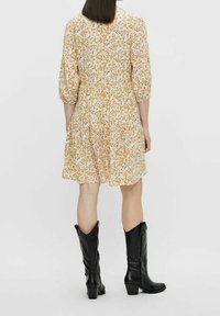Blumenmuster, kurzes Kleid mit kurzen Ärmeln in Creme mit orangefarbenen und blauen Akzenten; ausgestellter Rock und Knopfverschluss auf der Rückseite; kombiniert mit knielangen schwarzen Stiefeln.