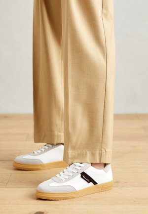 Beige pants worn with white and light gray lace-up sneakers featuring gum soles on a light wooden floor.