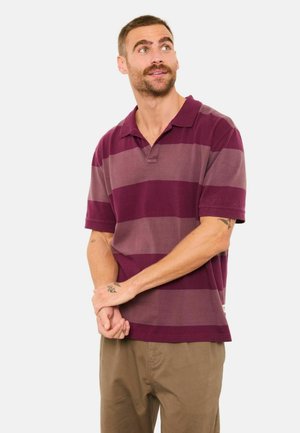 Bordeauxrotes und hellviolett gestreiftes Poloshirt, kurze Ärmel, aus Baumwollmaterial, mit lockerem Schnitt und einem Kragen. Es werden hellbraune Hosen getragen.