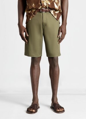 Uomo che indossa pantaloncini verde oliva fino al ginocchio, cintura marrone, camicia marrone a fiori e sandali intrecciati marroni, in piedi contro uno sfondo bianco.