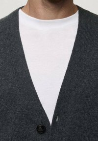 Dunkelgraue Strickjacke mit V-Ausschnitt, ausgestattet mit einem einzelnen schwarzen Knopf und über einem schlichten weißen T-Shirt getragen. Weiche Textur.