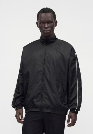 Homme portant une veste noire zippée avec un liseré blanc le long des manches, debout devant un fond clair uni.
