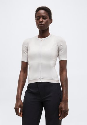 Γυναίκα με κοντά μαλλιά που φοράει εφαρμοστή ανοιχτογκρι μπλούζα με φερμουάρ και κοντά μανίκια, καθώς και μαύρο παντελόνι, στέκεται μπροστά σε απλό λευκό φόντο.