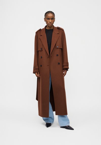 Γυναίκα με πλεξούδες, φοράει μακρύ καφέ διπλό παλτό, μαύρο πουκάμισο, ανοιχτό μπλε τζιν και μαύρα μυτερά παπούτσια, στέκεται με πρόσωπο προς τα εμπρός.