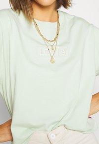 Světlezelené bavlněné tričko s vyšívaným logem "LEVI'S", doplněné o vrstvené zlaté řetízky. Uvolněný střih, krátké rukávy.