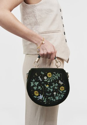 Person i beige outfit, der holder en sort håndtaske med broderede motiver af solsikker, blade, fugl, sommerfugl og halvmåne.