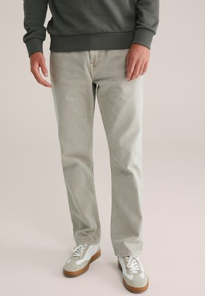 Man in een lichtgrijze rechte broek, witte en beige sneakers, en een donkergroen langmouwtop, staande op een effen achtergrond.