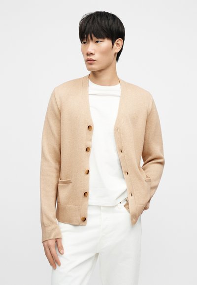 Ανοιχτό μπεζ ζακέτα, πλεκτή υφή, σχέδιο με V λαιμόκοψη, έξι καφέ κουμπιά, δύο εμπρόσθιες τσέπες, φορεμένη πάνω από λευκό πουκάμισο, στυλιζαρισμένη με λευκό παντελόνι.