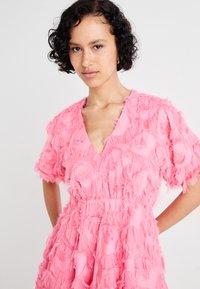 Ροζ φόρεμα με υφή και φτερωτό σχέδιο, σε σχήμα V στο ντεκολτέ και κοντά μανίκια, με σφιχτό τμήμα στη μέση για σιλουέτα που φαρδαίνει.