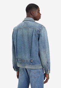 Un tânăr îmbrăcat într-o jachetă din denim de culoare albastru deschis și blugi, stând cu spatele parțial întors, pe un fundal alb simplu.