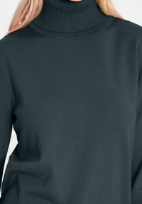 Pull-over à col roulé vert foncé en tissu côtelé. Présente une coupe ajustée avec un col montant et des manches longues.