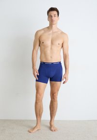 En mann uten skjorte som står barbeint på et teppebelegg iført blå Björn Borg boxershorts foran en enkel hvit vegg.