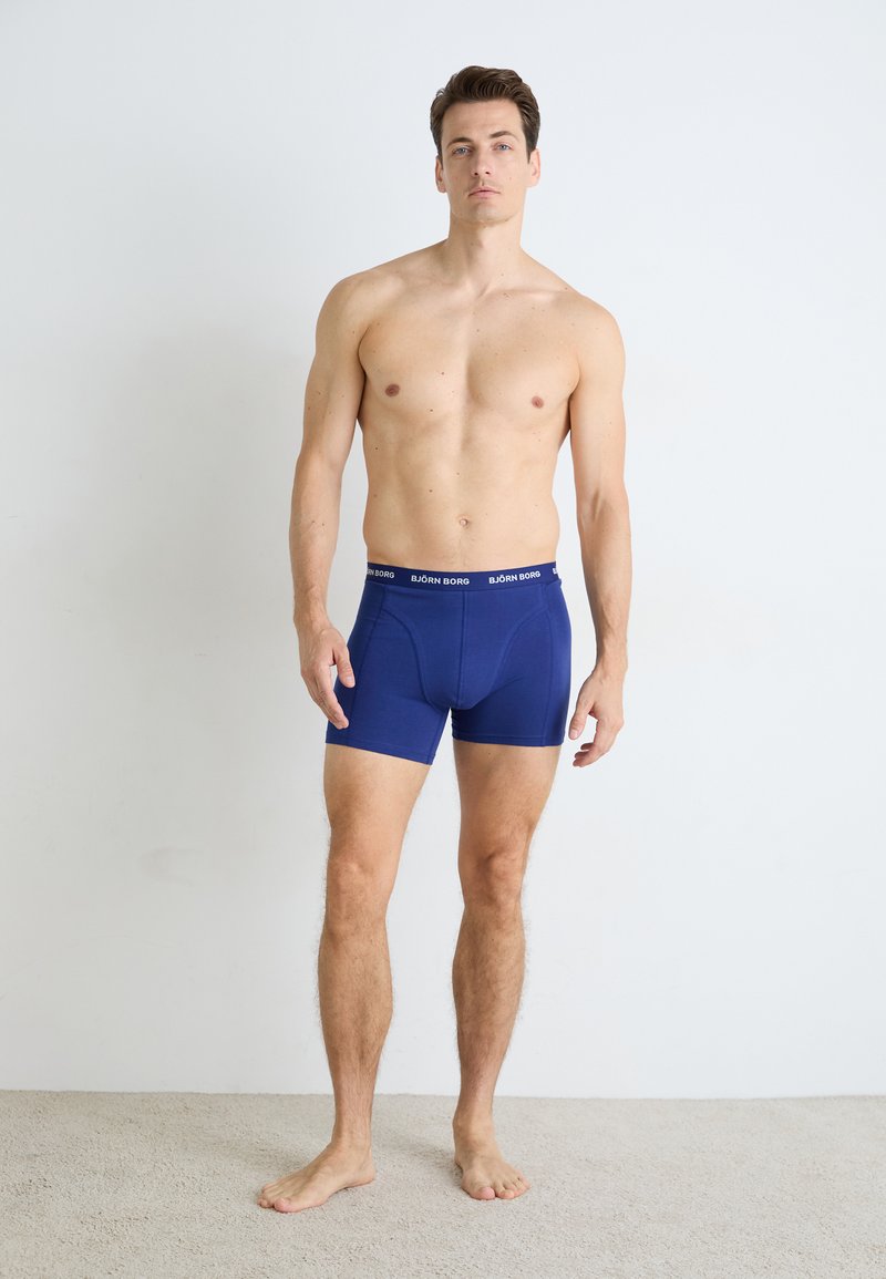 Felsőtest nélküli férfi mezítláb áll egy szőnyeggel borított padlón, kék Björn Borg boxer alsót viselve, egy sima fehér fal háttérében.