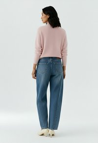 Femme debout, de dos, portant un pull rose clair, un jean bleu ample et des chaussures blanches à talons, sur un fond uni clair.