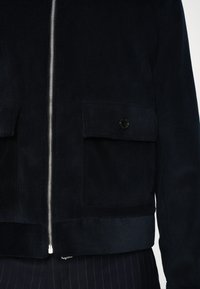 Navy corduroy jacket with a front zip, two large patch pockets, and a ribbed hem. Features a logo button and textured vertical lines.