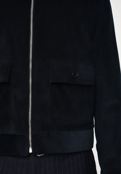 Veste en velours côtelé marine avec une fermeture éclair à l'avant, deux grandes poches appliquées et un ourlet côtelé. Dispose d'un bouton logo et de lignes verticales texturées.