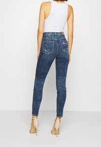Jeans ajustados de mezclilla azul oscura, con cintura alta, bolsillos traseros, detalles desgastados y un diseño de pierna entallada. Combinados con tacones beige.