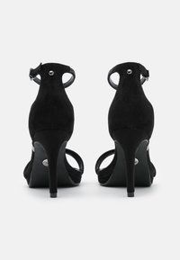 Sandali alti in suede nero con un sottile cinturino attorno alla caviglia, un bordo scallopato texturizzato e un accent metallic nel tacco.