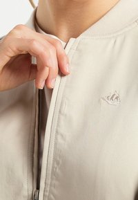 Cazadora bomber de color beige claro con una textura suave, cuello acanalado y detalles de cremallera, que presenta un pequeño logo bordado en el pecho.