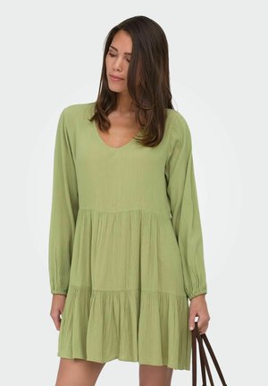 Femme portant une robe ample à manches longues de couleur vert clair, tenant des poignées brunes de sac à main, regardant vers le bas, yeux fermés.
