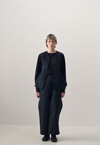 Zwarte vest met meerdere zakken over een zwart longsleeve top, gecombineerd met zwarte wijde broek en zwarte schoenen. Eenvoudig ontwerp en textuur.