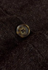 Nagyított kép egy barna márványozott, négylyukú gombáról, amely sötétbarna kötött anyagra van varrva, látható textúrával és szálakkal.