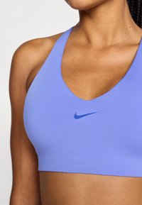 Μωβ αθλητικό σουτιέν με βαθύ V λαιμόκοψη, κατασκευασμένο από λείο ύφασμα, με μικρό μπλε λογότυπο της Nike στο κέντρο.