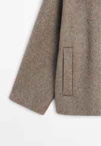 Brązowy wełniany płaszcz o fakturowanym wykończeniu, szerokich rękawach i minimalistycznym detalu kieszeni naszywanej po boku.