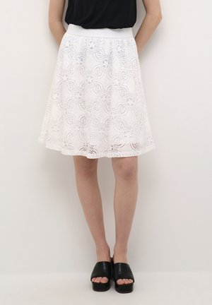 Femme portant une jupe blanche en dentelle longueur genoux avec des motifs floraux, un haut noir et des chaussures à plateforme noires à bouts ouverts, debout devant un fond uni.