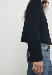 Svart texturerad kort jacka med rund krage, med framfickor och tre knappdekorationer på ärmarna, i kombination med blå denimjeans.