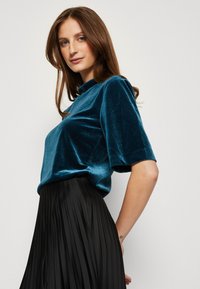 Teal fluwelen top met korte mouwen en hoge hals, gecombineerd met een zwarte geplooide rok. Glad textuur, vloeiende uitstraling, en een aansluitende silhoette.