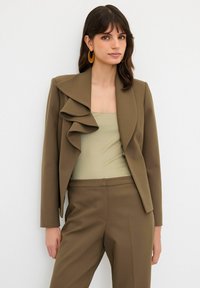 Hnědý upravený blazer s volánkovým detailem na límci, spárovaný se světle zeleným vypasovaným topem a odpovídajícími hnědými kalhotami. Hladká textura.