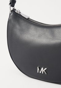 Borsa a mano in pelle nera con un design curvo, hardware in argento e logo "MK" in rilievo. Presenta una cerniera metallica lungo il bordo superiore.