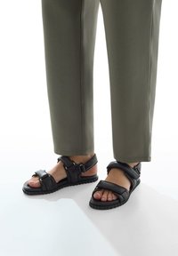Zwarte leren sandalen met verstelbare banden, een gemarmerde rubberen zool, blootgestelde tenen en accenten van goudkleurig hardware, gedragen met olijfgroene broek.