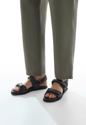 Zwarte leren sandalen met verstelbare banden, een gemarmerde rubberen zool, blootgestelde tenen en accenten van goudkleurig hardware, gedragen met olijfgroene broek.