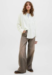 Camicia button-up crema con tasca, abbinata a jeans in denim marrone a gamba larga. Tessuto morbido con taglio rilassato, caratterizzato da una finitura sbiadita.