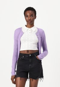 Cárdigan de punto lavanda con diseño calado, combinado con una blusa corta blanca que presenta un patrón texturizado y un cuello, y pantalones cortos de mezclilla negros.