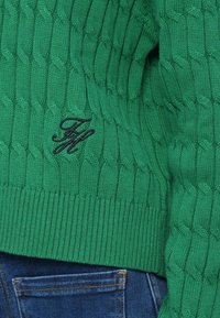 Zöld kötött pulóver, texturált kábelmintával, bordás mandzsettával és szegéllyel, bal oldalon sötétkék hímzett logóval.
