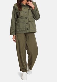 Olijfgroene jas met vier voorzakken, knoopsluiting en lange mouwen, gecombineerd met loose-fit olijfkleurige broek en witte sneakers.