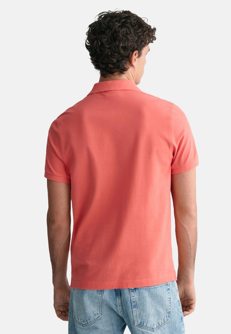 Mann mit lockigem Haar, von hinten gesehen, trägt ein korallenfarbenes Poloshirt mit kurzen Ärmeln und hellblaue Jeans vor einem einfarbigen Hintergrund.