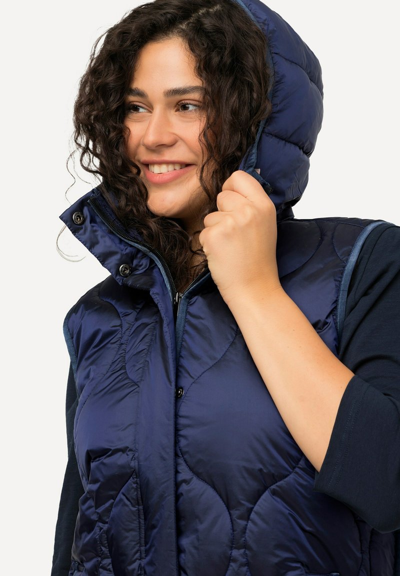 Marineblaue Steppweste mit einem klassischen Muster, hohem Kragen und Kapuze. Verfügt über Druckknöpfe und einen Reißverschluss vorne. Strukturierter Stoff.