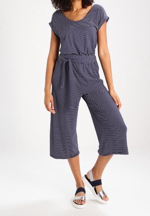 Nainen pukeutuneena laivastonsiniset ja valkoiset raidalliset lyhythihaiset haalariin, joissa on leveät lyhyet housut, sekä hopeiset sandaalit, joissa on laivastonsiniset remmit.