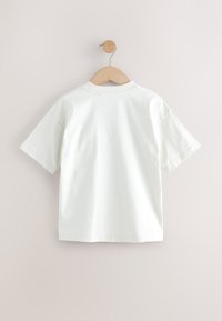 Tricou alb din bumbac, cu mâneci scurte, gât rotund și croială lejeră. Atârnă pe un umeraș din lemn, pe un fundal deschis la culoare.