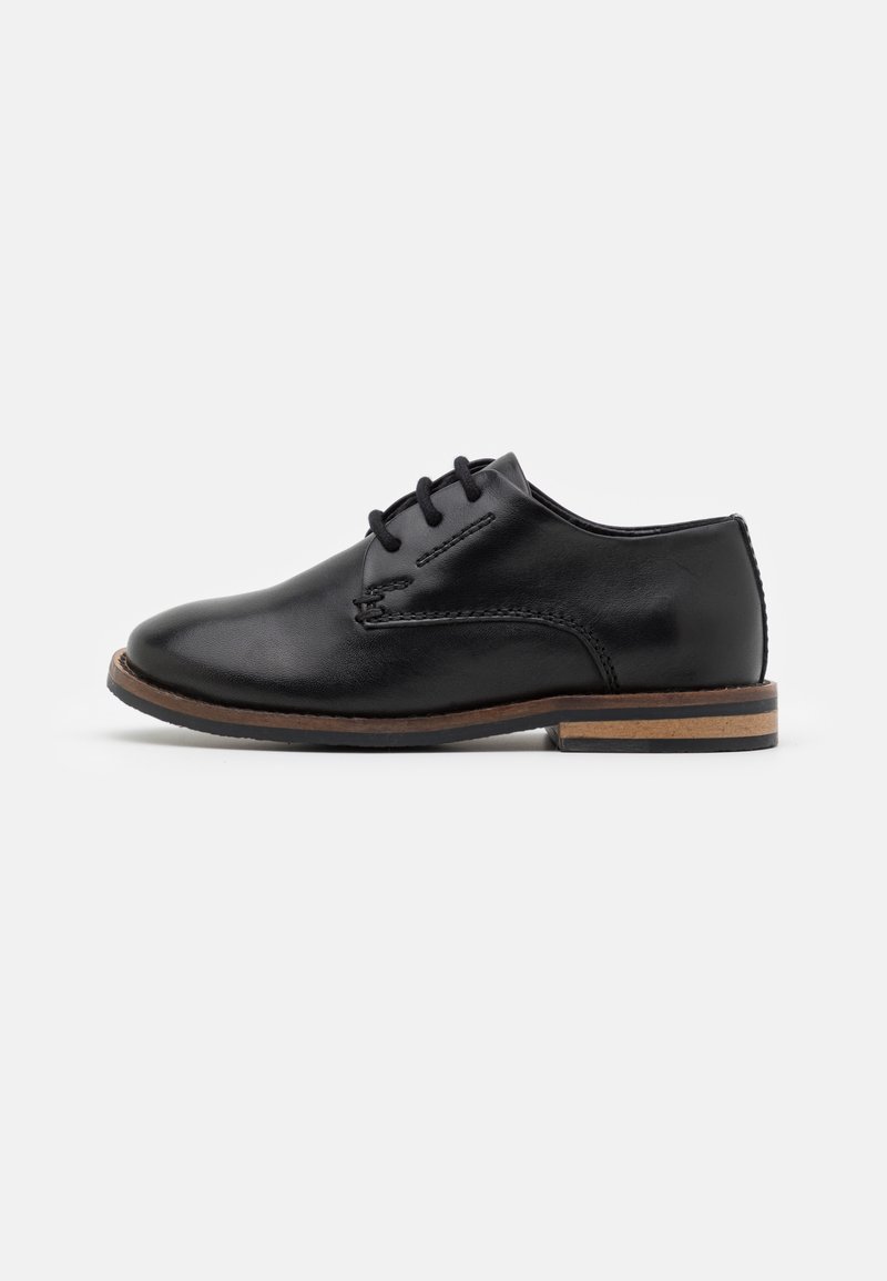 Black leather shoe with a smooth texture, minimalistic design, rounded toe, four eyelets, and a contrasting brown sole accent.