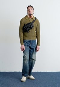 Oliwkowy sweter z dzianiny z kołnierzykiem, niebieskie dżinsy, białe trampki i czarna torba na ramię z plecionym wzorem. Prosta, casualowa stylizacja.